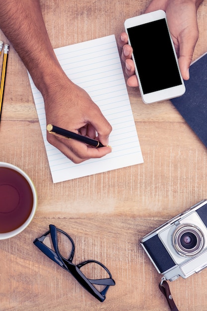 High angle view of businessman holding smart phone while writing on notepad by camera and coffee on table