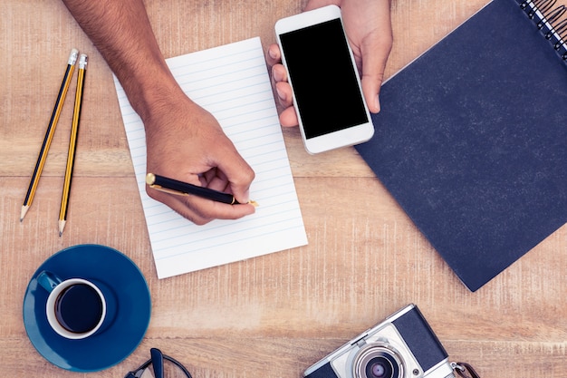 High angle view of businessman writing on notepad while holding smart phone by camera and eyeglasses on table