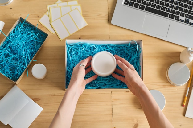 High angle view of female worker putting cream in cardboard box at table for delivery