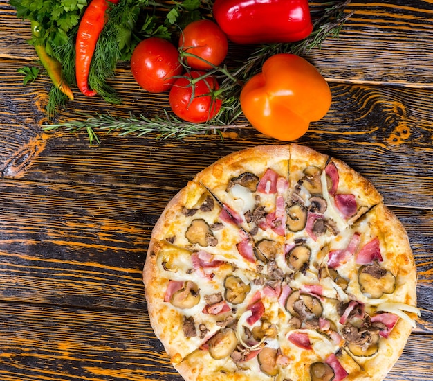 High angle view of pizza with ham, mushrooms, onion and pickles near are peppers, tomatoes, sprig of rosemary  and other vegetables on wooden table