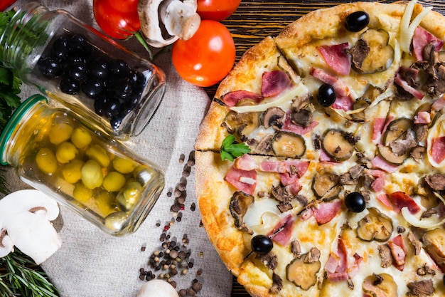 High angle view of pizza with ham, onion, black olives on wooden table, near lie on a napkin tomatoes, mushrooms and jars of pickled vegetables