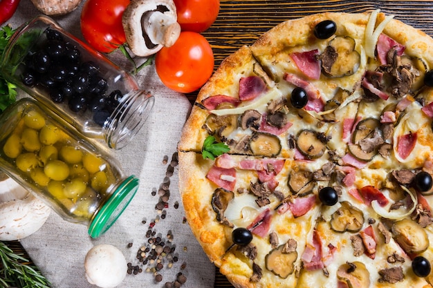 High angle view of tasty pizza with ham, onion, black olives on wooden table, near lie on a napkin tomatoes, mushrooms and jars of pickled vegetables