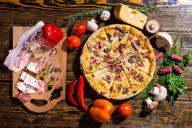 High angle view of tasty pizza on wooden table surrounded by vegetables, mushrooms and cheese near cutting board with meat