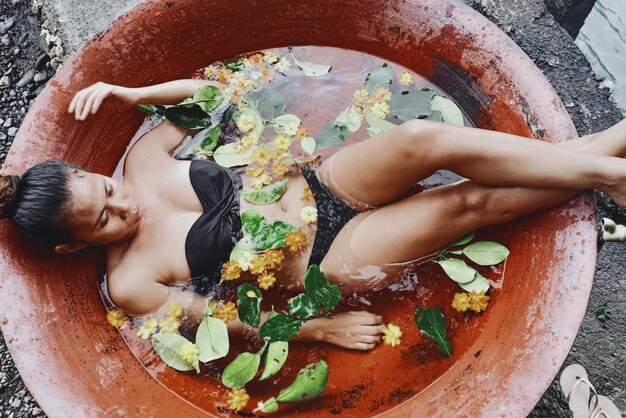 Photo high angle view of woman wearing bikini while lying in container with water and leaves