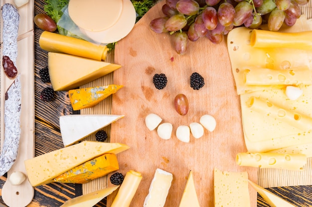 Photo high angle view of wooden cutting board surrounded by variety of cheeses and happy face made from bocconcini and blackberries