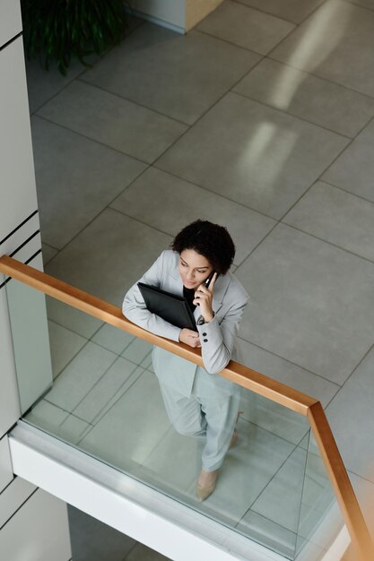 High angle view of young businesswoman in suit talking on mobile phone standing on balcony of office