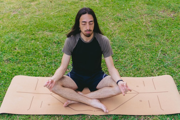 High angle of a young Hispanic and Latino hippie male meditating with his eyes closed while doing the lotus pose on a yoga mat in a pasture