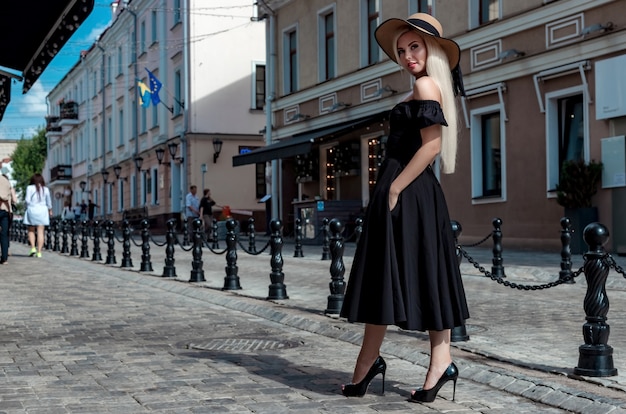 High fashion portrait of a graceful woman in an elegant straw hat and dress that walks along a city street