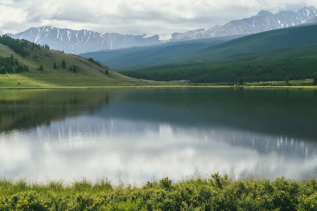 High mountains in cloudy sky and lake near green grasses and forest hills.