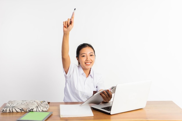 High school girl at desk holding book with pointing finger while using laptop on isolated background