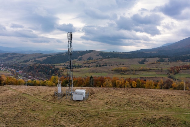 High steel cell tower near settlement on mountain top covered with autumn forest under gloomy sky with thick grey clouds panorama view