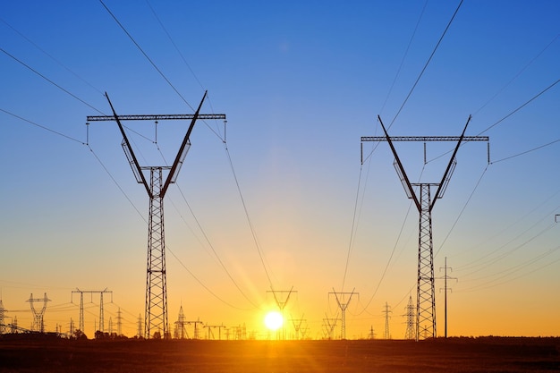 High voltage electricity towers in field at sunset and clear blue sky Dark silhouettes of repeating power lines on orange sunrise Electricity generation transmission and distribution network