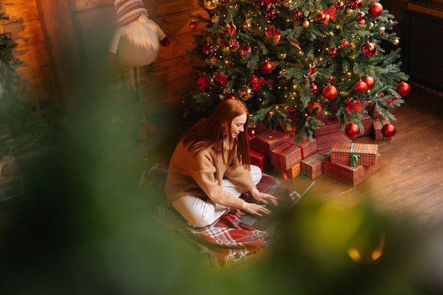 Highangle shot of happy young redhead woman wearing winter sweater typing online message on laptop