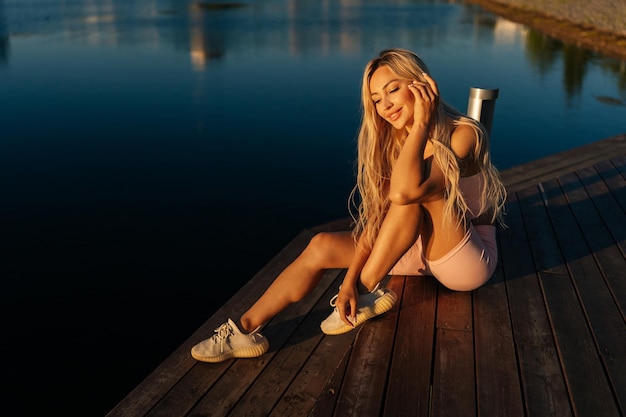 Highangle view of cheerful sporty blonde woman in sportswear sitting posing on wooden pier cite park by water on sunny summer morning looking down