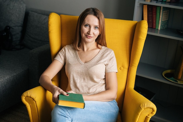 Highangle view of pretty blonde woman resting in comfortable yellow chair holding paper book in dark room with modern interior