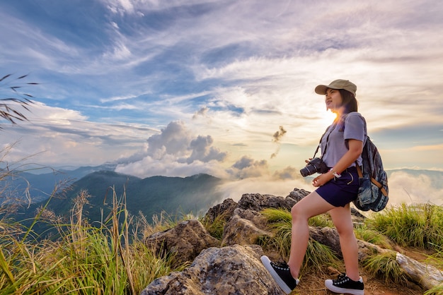 Hiker Asian cute teens girl happy with camera backpack and cap looking beautiful nature landscape sky at sunset on mountain, Adventure travel in Asia at Phu Chi Fa Forest Park, Chiang Rai, Thailand