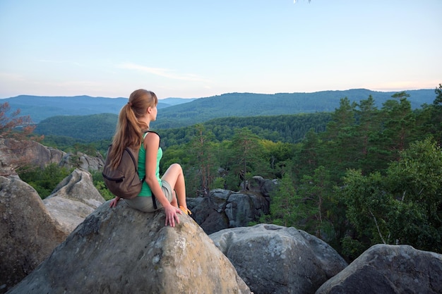 Hiker girl resting on rocky mountain top enjoying morning nature during her travel on wilderness trail Lonely female traveler traversing high hilltop route Healthy lifestyle concept