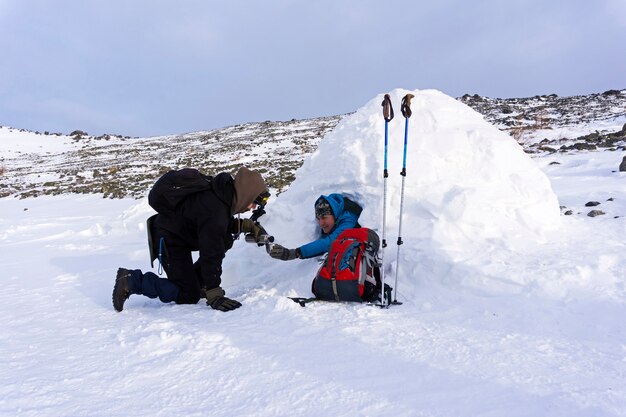 Hiker pours from the thermos tea to his friend sitting in a snowy house igloo