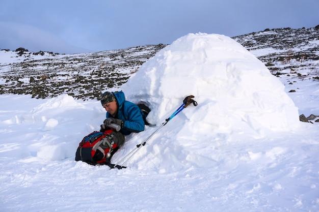 Hiker pours himself a hot drink from a thermos sitting in a snowy house igloo