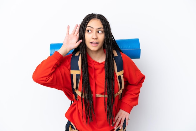 Hiker teenager girl with braids over isolated white background listening to something by putting hand on the ear