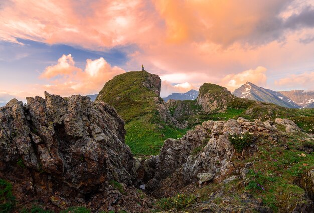 Hiker with a backpack on a green mountain peak against the backdrop of the sunset sky