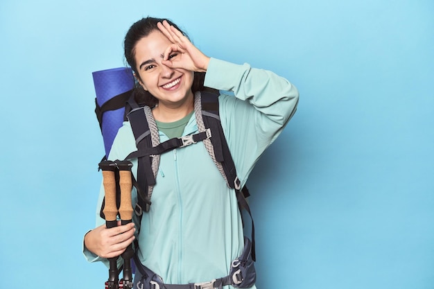 Hiker with backpack and poles on blue excited keeping ok gesture on eye