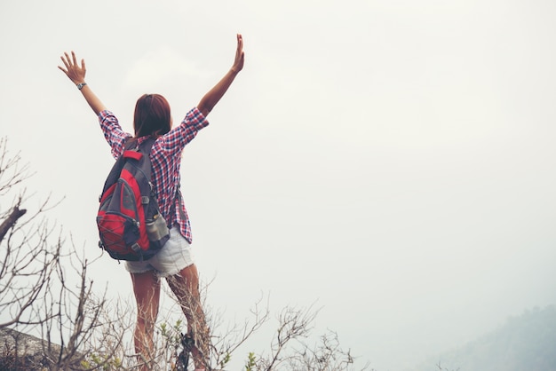 Hiker with backpack standing on top of a mountain with raised hands and enjoying nature view.