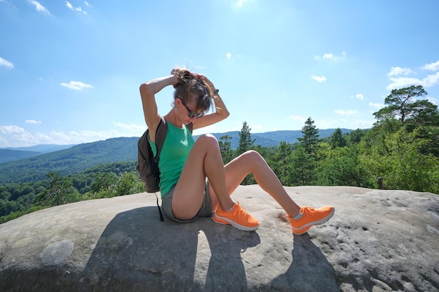 Hiker woman resting on rocky mountain top enjoying nature during her travel on wilderness trail Lonely female traveler traversing high hilltop route on hot summer day Healthy lifestyle concept