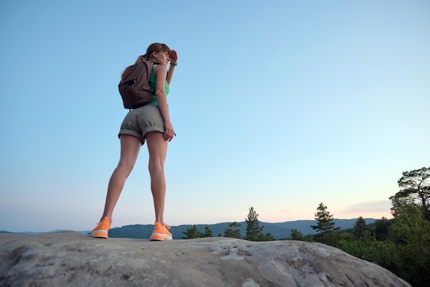 Hiker woman standing on mountain footpath enjoying evening nature during her travel on wilderness trail Lonely female traveler traversing high hilltop route Healthy lifestyle concept