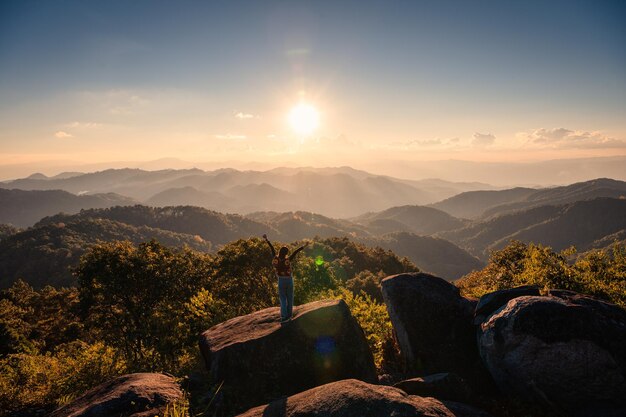 Hiker woman standing and raising arms on rock with sunset over horizon in tropical rainforest at national park Doi Kham Fah viewpoint Wiang Haeng Chiang Mai Thailand