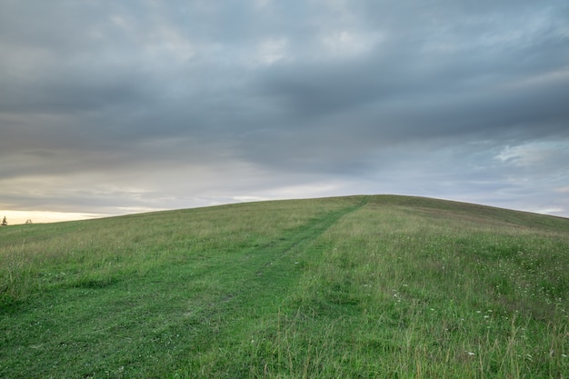 Hill with green grass and pathway against the sky with storm clouds at sunset