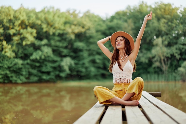 Hippie ecoactivist woman traveler sits on a bridge by a lake with her arms outstretched with a hat and smiling sincerely