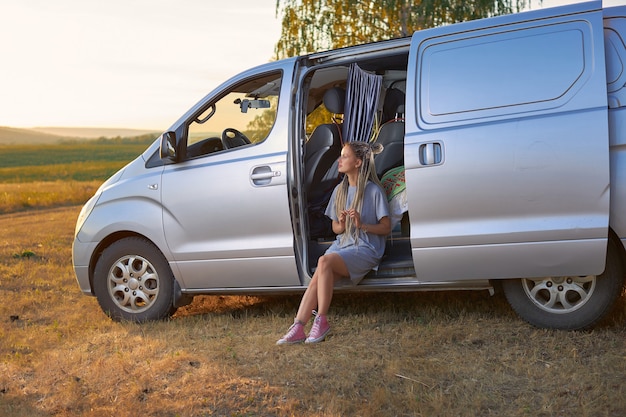 A hippie girl sits in a silver minivan on a field against the backdrop of mountains the concept of f...