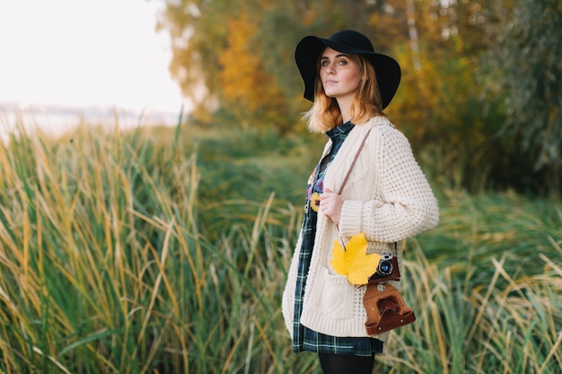 Hippie girl with a yellow maple leaf in a knitted sweater and hat walks autumn park.