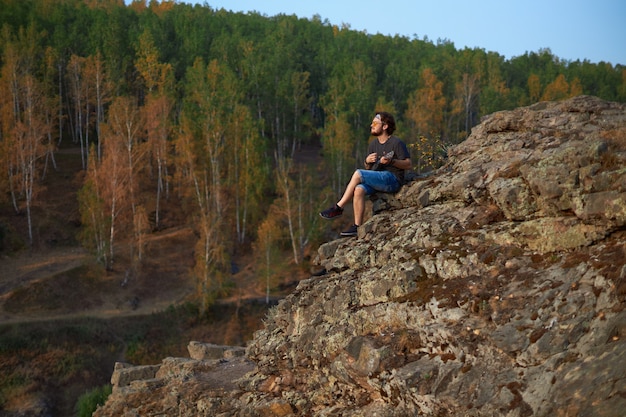 A hippie guy in glasses and denim shorts plays the ukulele sitting on top of a cliff in the middle o...