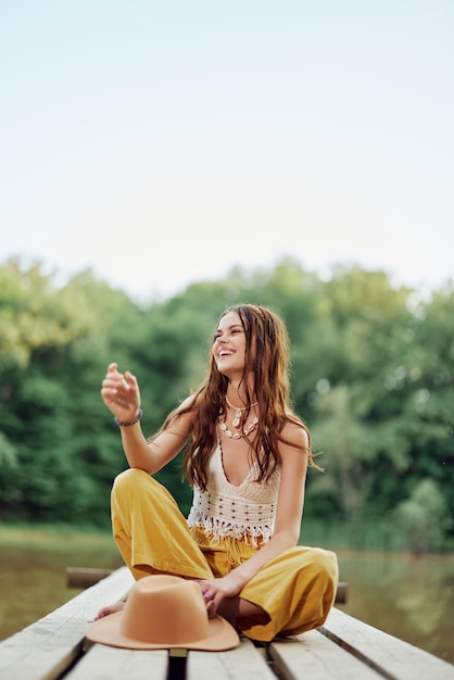 A hippie woman sits on a bridge by a lake on a nature trip and smiles in ecoclothing The concept of ecoactivist and nature conservation High quality photo