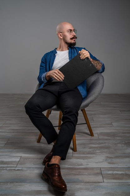 Hipster bearded man with moustache wearing glasses who sitting on chair in studio isolated grey back