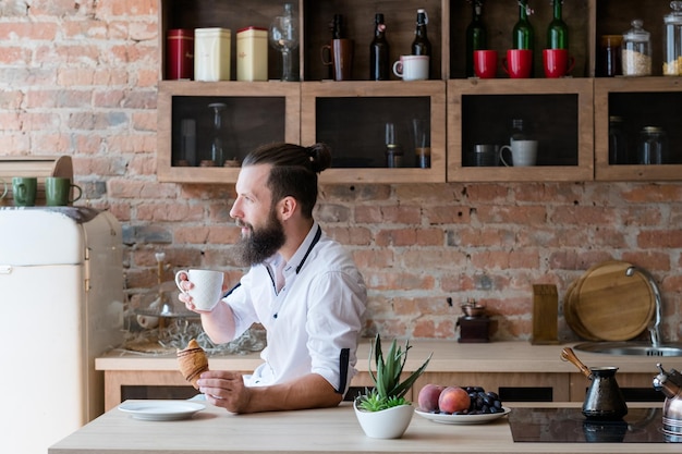 Hipster breakfast New day Contemplation Bearded man with cup of coffee and croissant looking sideways Loft apartment