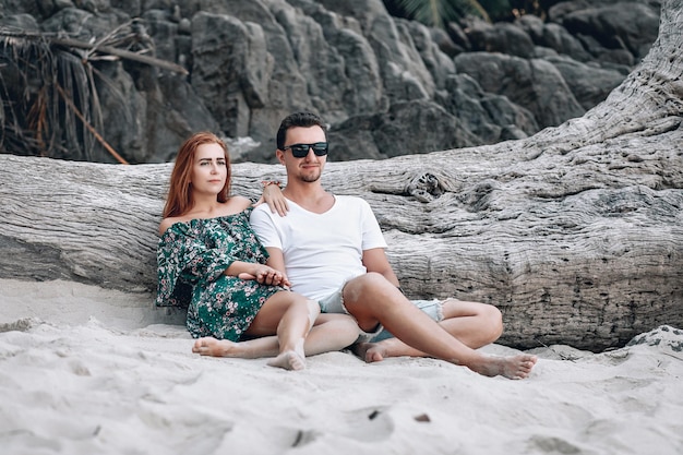 Hipster couple on the beach, girl with red hair in green floral dress and man with stubble, sitting near old big bare tree without leaves. Phuket. Thailand