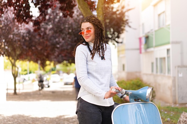 Hipster girl in pink sunglasses with dreadlocks standing near vintage motor scooter on city street