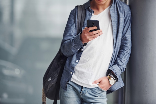 Hipster guy in nice clothes stands indoors with phone in hand