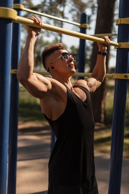 Hipster handsome muscular sports man with headphones and sunglasses in a black tank top pulls himself up on the bar and workout in the park healthy lifestyle