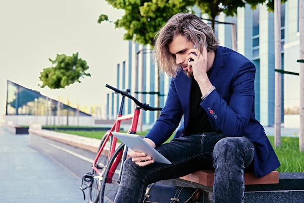 Hipster male with long blond hair talks by smart phone and holds tablet PC with single speed bicycle on background.