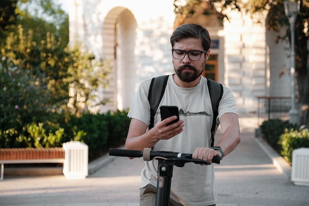 Hipster man commuter with electric scooter outdoors in city using smartphone