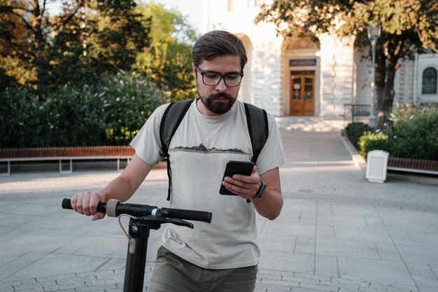 Hipster man commuter with electric scooter outdoors in city using smartphone