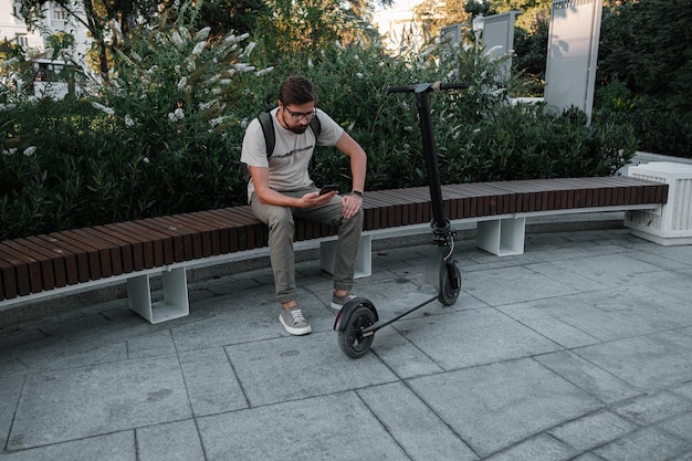 Hipster man commuter with electric scooter outdoors in city using smartphone