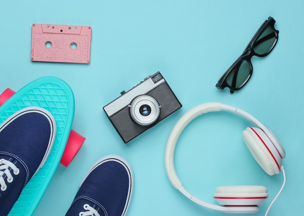 Hipster outfit. Skateboard with headphones, audio cassette, retro camera and sneakers on blue wall