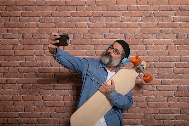 A hipster takes a selfie on his smartphone with the longboard on a brick wall.