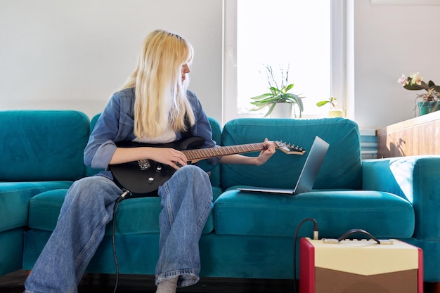 Hipster teenager girl playing electric guitar looking at laptop monitor