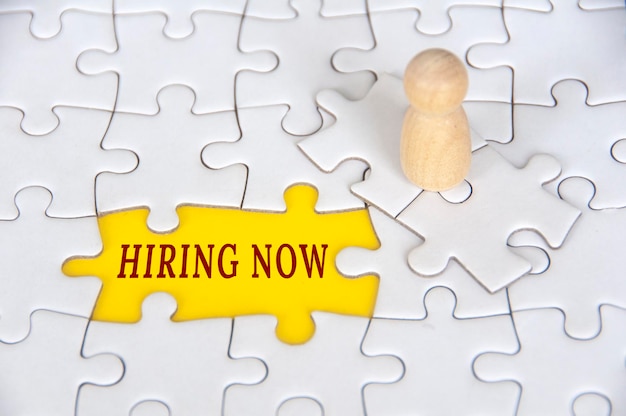 Photo hiring now text on missing jigsaw puzzle with wooden figure background employment concept